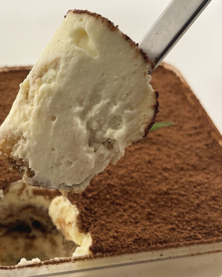 Close-up of a spoon lifting tiramisu out of the tub: mascarpone cream and a coffee-soaked ladyfinger under the cocoa
